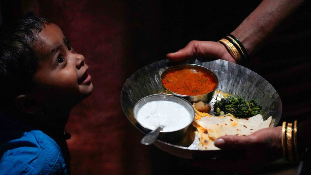 A young Indian girl is offered a plate of food by her mother inside their home in India. 