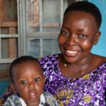 A Sierra Leonean mom sits smiling with her toddler son on her lap.