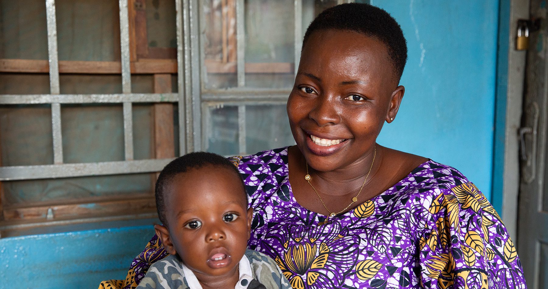 A Sierra Leonean mom sits smiling with her toddler son on her lap.