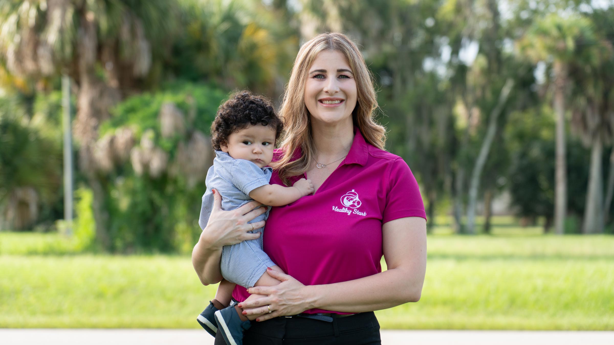Andrea, Executive Director of Okeechobee Healthy Start Coalition, stands smiling and holding her infant son.