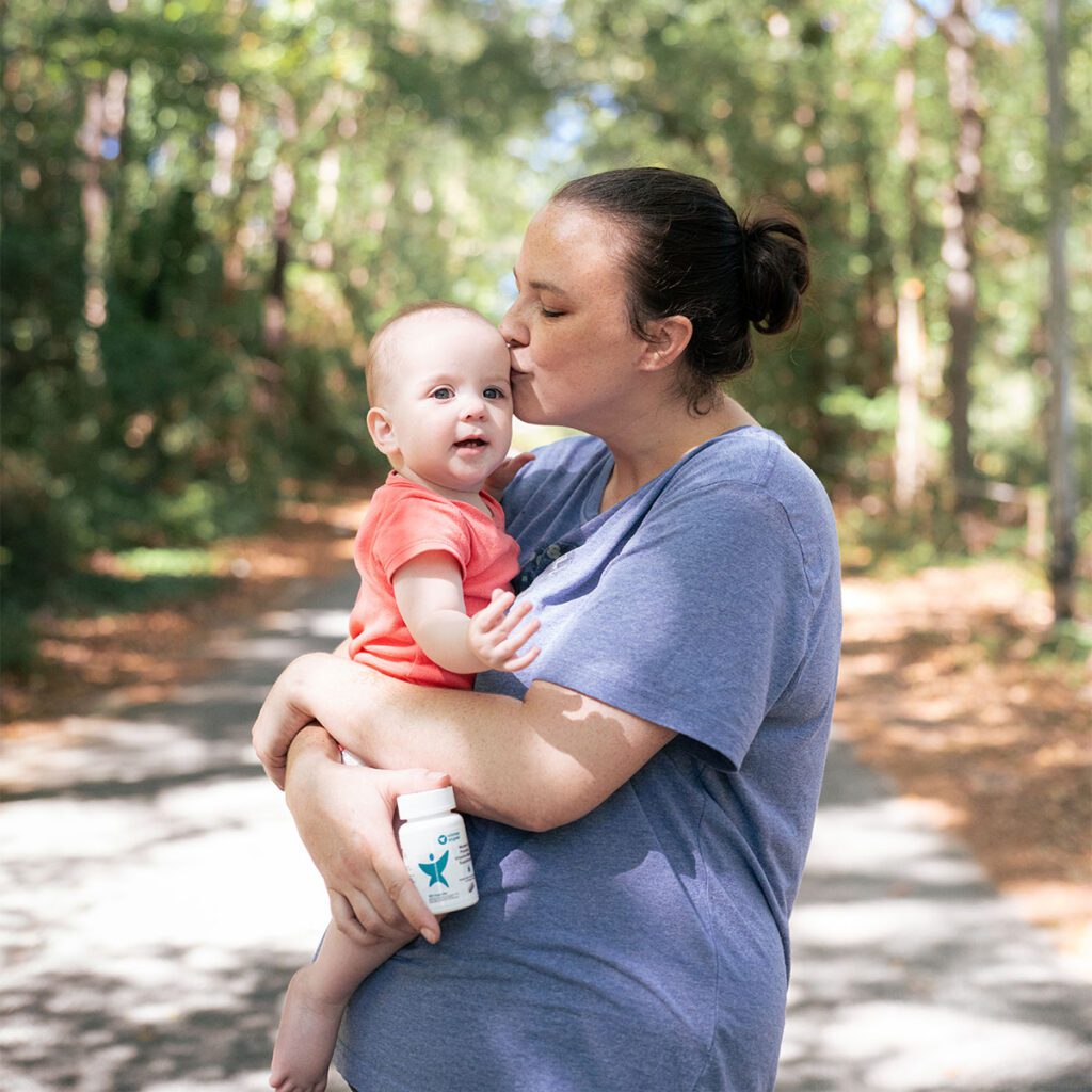 Kaylene holding and kissing infant baby on the head while holding a bottle of prenatal vitamins and minerials.