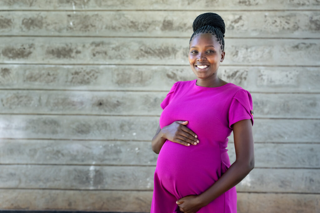 Portrait of a smiling woman with her arms framing her pregnant midsection in Kenya.