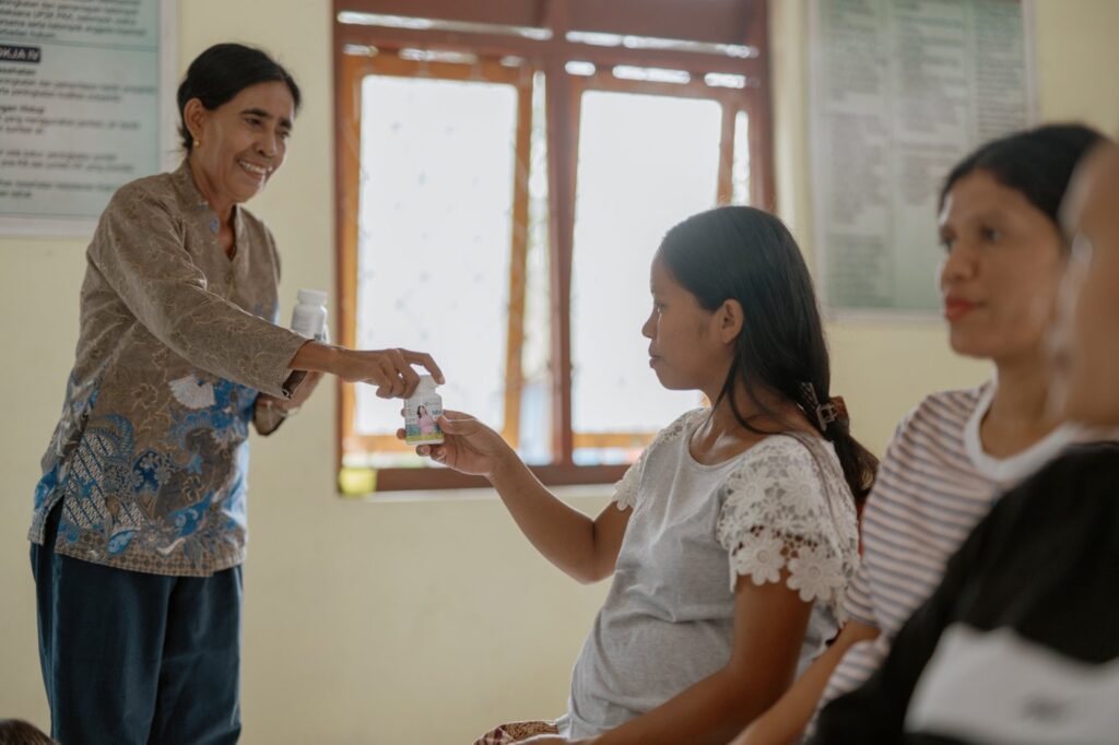 A smiling health worker hands a bottle of prenatal multiple micronutrient supplements to a pregnant woman seated in a clinic.
